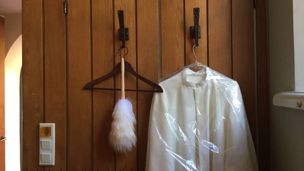 Holzwand mit zwei Haken, daran ein Staubwedel und weiße Gewänder in Plastikhülle.