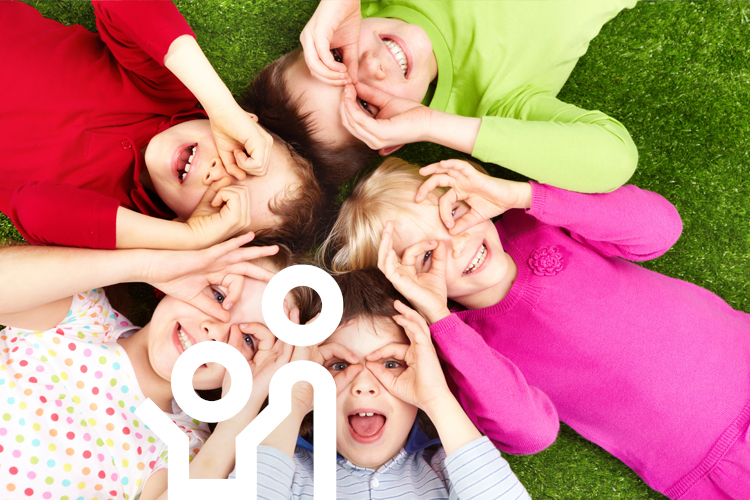 Fünf Kinder liegen lachend im Kreis auf einer grünen Wiese und formen mit ihren Händen spielerisch Ferngläser vor den Augen.